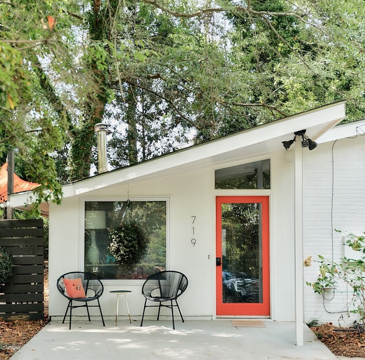 The white mid-century house under its trees: slanted roof, orange front door and two butterfly chairs on the front patio