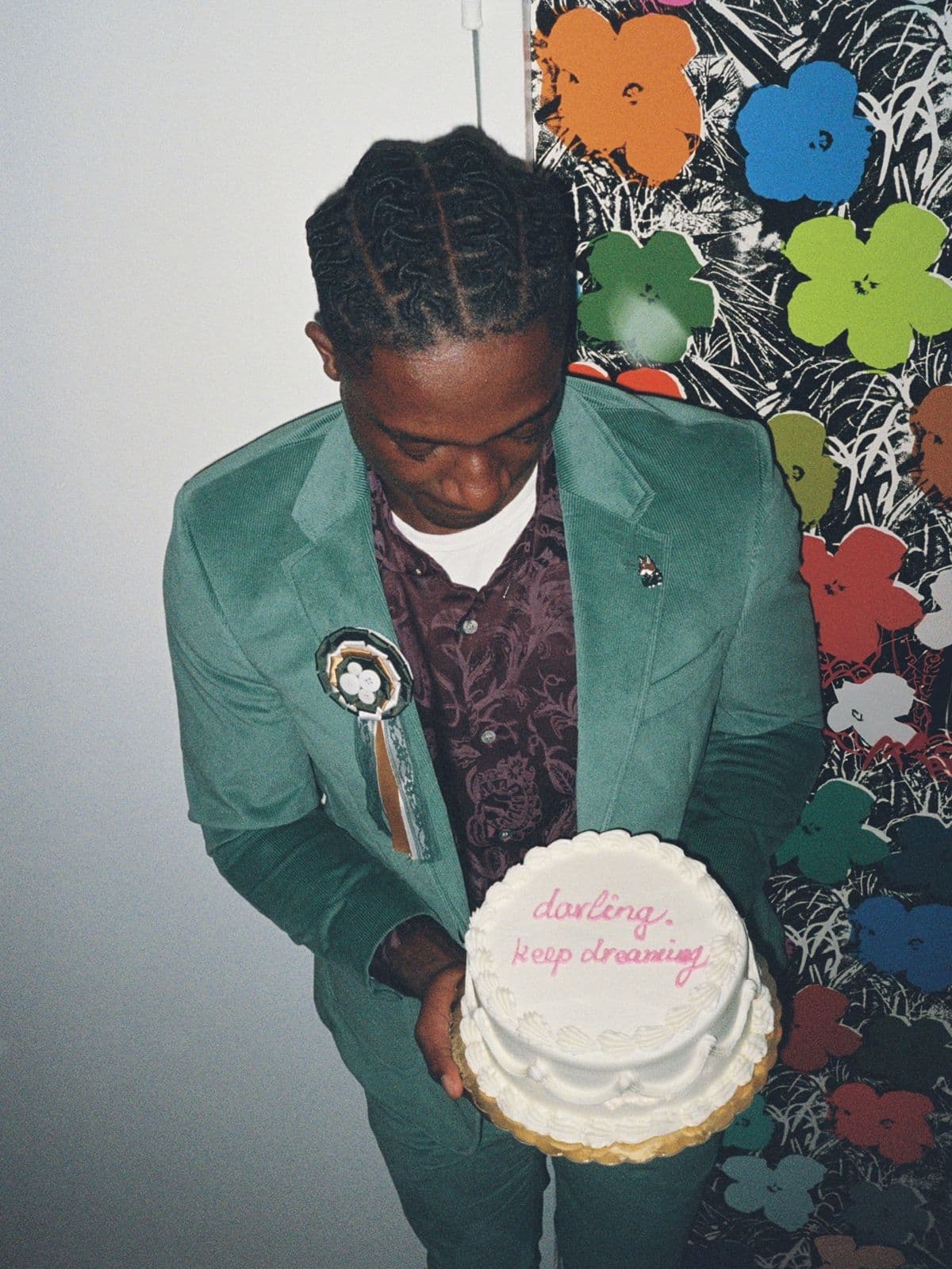 Man in a green suit holding a frosted cake in front of the flower print wallpaper